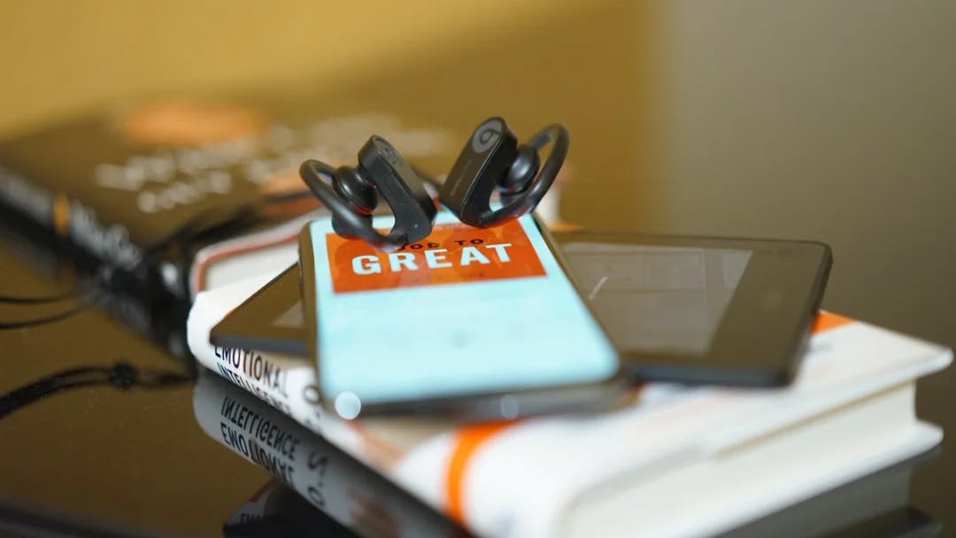 Lydbok på mobilen med ørepropper. Ligger på toppen av en bok. Foto.